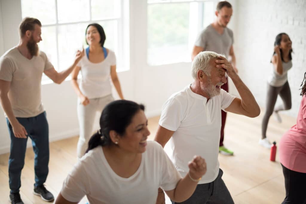 Groupe diversifié de personnes souriantes, dont un homme senior riant, s'exerçant avec énergie dans un cours de fitness lumineux.