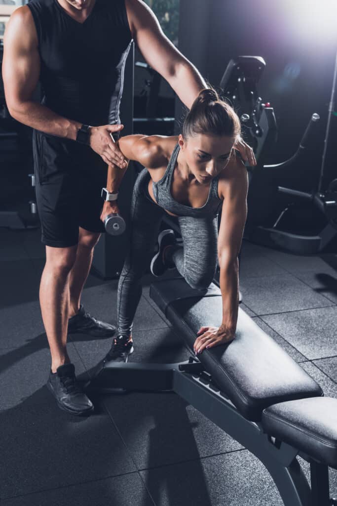 Tirage Haltère : Femme S'entraîne en Salle avec Coach Pro Une femme concentrée s'entraîne avec un haltère (4kg) sur un banc en salle de sport, aidée par son coach personnel.