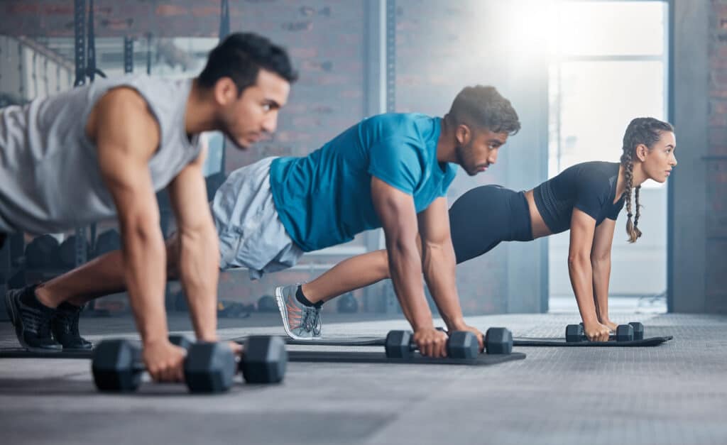 Trois personnes s'entraînent avec des haltères dans une salle de sport, effectuant des pompes ou des planches. Ils sont concentrés sur l'exercice.