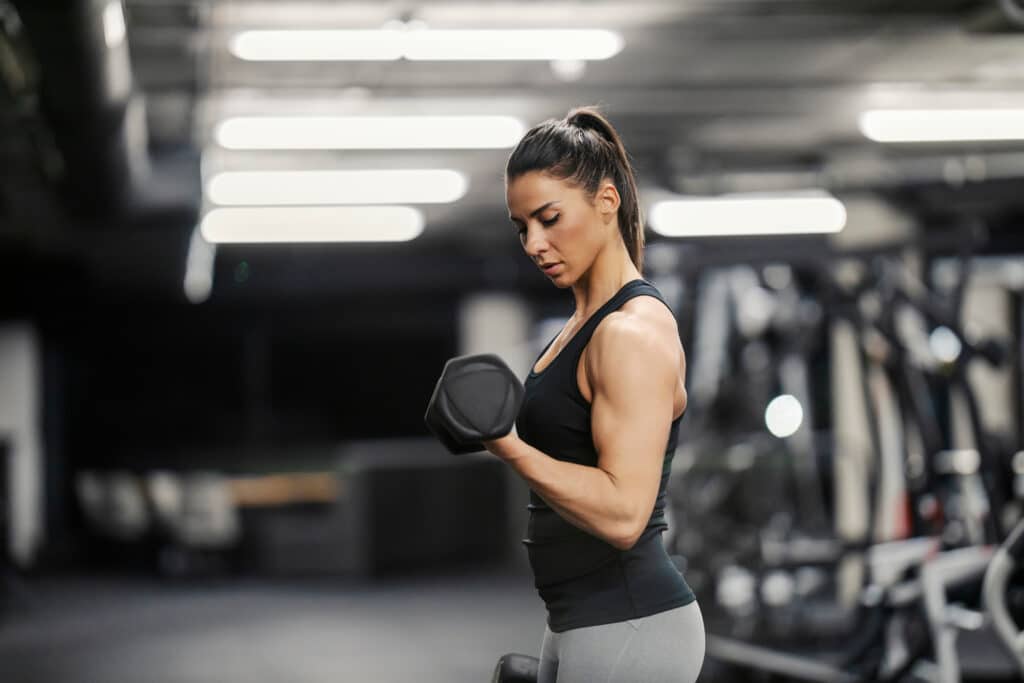 Femme concentrée s'entraînant dans une salle de gym, faisant des biceps curls avec un haltère noir. Elle porte un débardeur noir et un legging gris.
