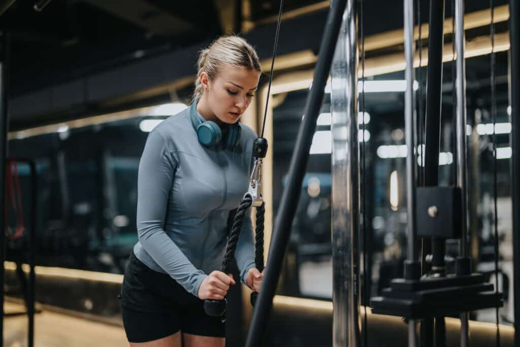 Femme s'entraîne à la musculation avec câble en salle de sport Jeune femme blonde concentrée s'entraîne sur machine à câble en salle de sport. Elle porte un haut bleu, short noir Nike et écouteurs bleus.