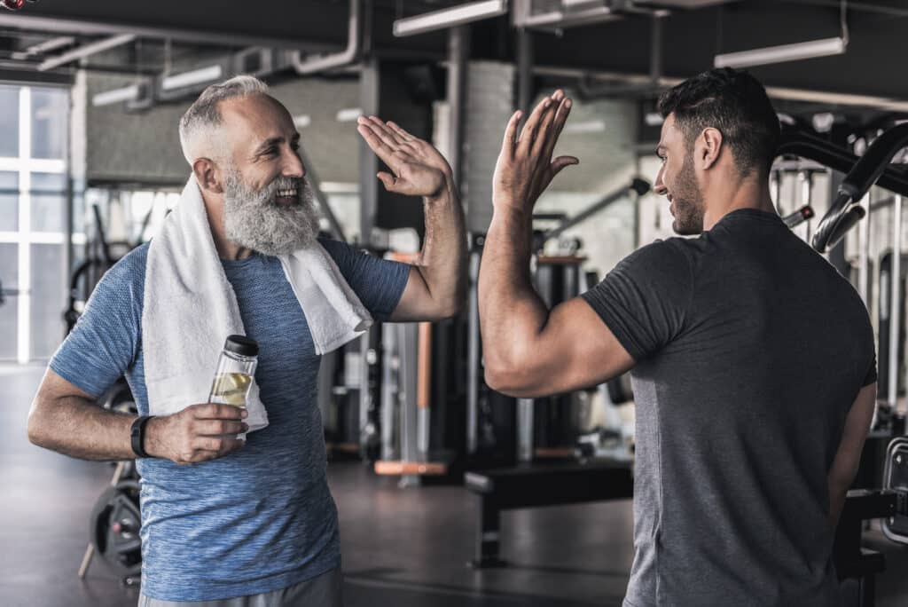 Deux hommes souriants se donnent un high-five dans une salle de sport. L'homme âgé barbu tient une bouteille d'eau.