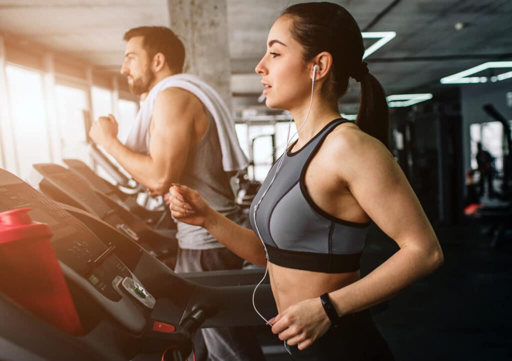 Une femme concentrée court sur un tapis de course dans une salle de sport, écoutant de la musique. Un homme s'entraîne derrière elle.