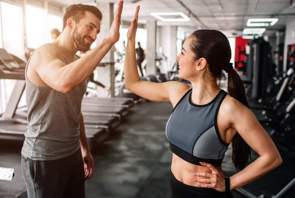 Deux amis souriants, un homme et une femme en tenue de sport, se donnent un high-five motivant dans une salle de sport moderne.