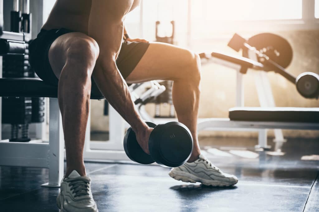 Homme musclé, torse nu en short, fait un exercice d'haltère sur un banc de gym lumineux. Il est concentré sur son entraînement de force.