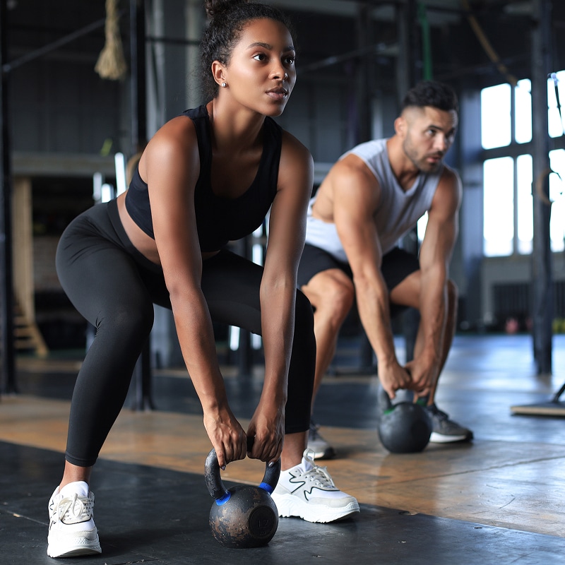 Femme et homme s'entraînent au gym, réalisant un soulevé de kettlebell. La femme, concentrée, tient une kettlebell.