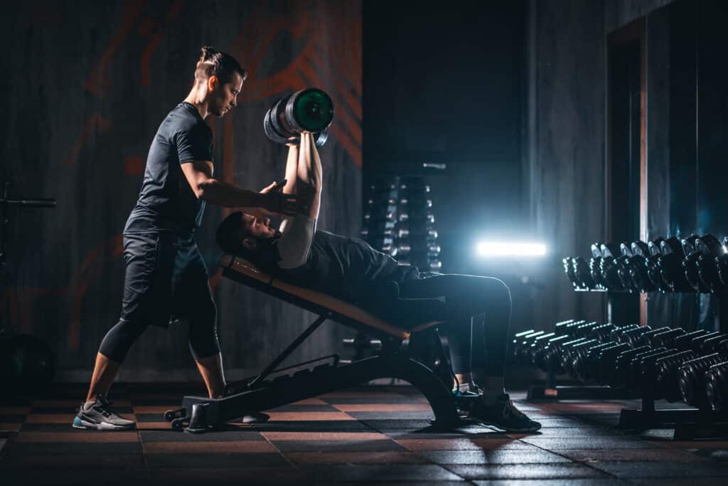 Homme soulevant un haltère de 26kg sur un banc incliné, spoté par son coach dans une salle de sport sombre. Effort intense.