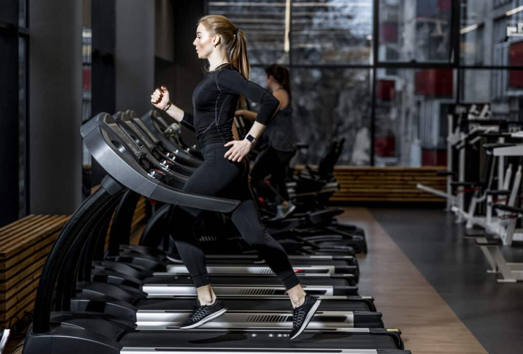 Sportive en tenue noire, courant sur un tapis de course moderne. Salle de sport lumineuse avec fenêtres et équipements.